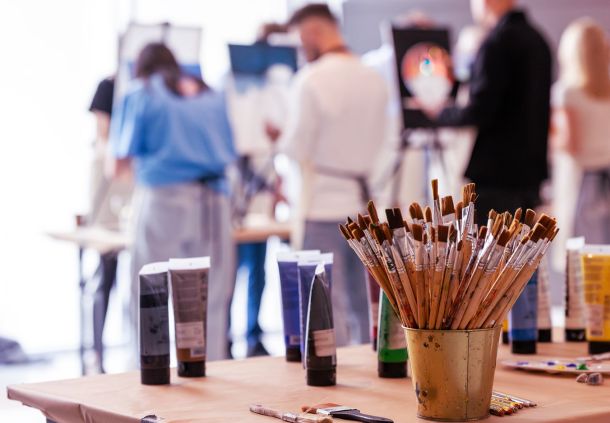 Tisch mit verschiedenen Pinseln und Farben, im Hintergrund malen Personen an Staffeleien